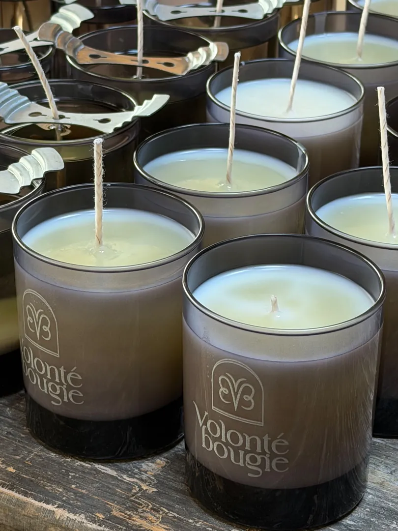 Velas recién vertidas en vasos ahumados con el logotipo de Volonté Bougie; unas pinzas metálicas centran las mechas